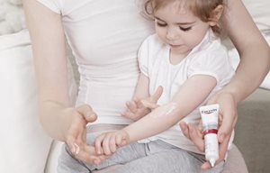 Holčička sedí u maminky na klíně a maže si Eucerin AtopiControl Acute krém na ohyb ruky.